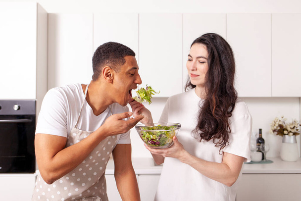 Mutfakta birlikte salata hazırlayan çok ırklı bir çift, Amerikalı erkek ve Avrupalı kız, beyaz modern mutfakta sebze ve sebzelerden sebze yiyor, mutlu, ırklar arası mutlu bir aile. - Fotoğraf, Görsel