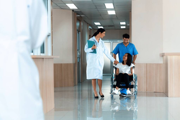Lääkäri ja mieshoitaja kuljettavat naispotilaan pyörätuolissa steriiliä sairaalakäytävää pitkin. Vammaisten terveydenhuolto ja sairaanhoito sairaalakonseptissa. - Valokuva, kuva