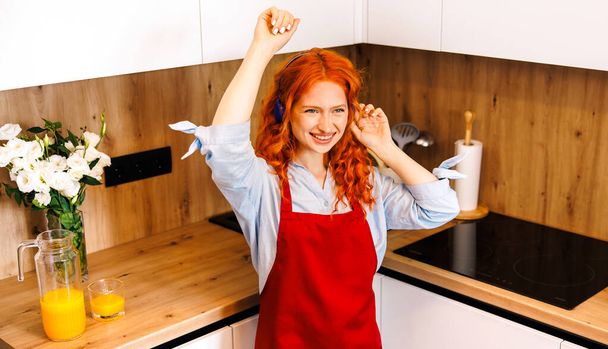 Fun and relax. Cooking and preparing meal in house kitchen. Funny cute housewife woman wearing earphones dancing with spoon ladle. Indoor. High quality photo - Foto, Imagem
