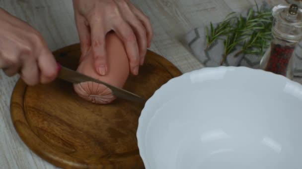 Cutting boiled sausage into circles with a kitchen knife on wooden cutting board. Close-up female hands slicing sausage on wooden cutting board with iron kitchen knife. Pepper grinder, rosemary sprig. - Video