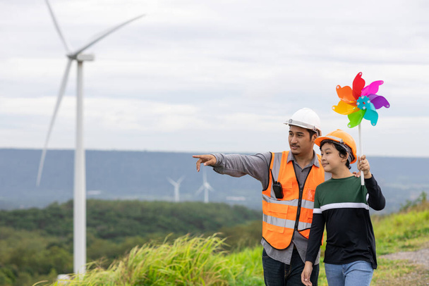 Engineer with his son holding windmill toy on a wind farm atop a hill or mountain. Progressive ideal for the future production of renewable, sustainable energy. Energy generated from wind turbine. - Foto, immagini