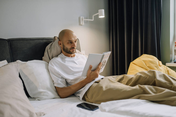 A man in home clothes is lying in a white bed in a bright bedroom reading a book in the afternoon - Foto, afbeelding