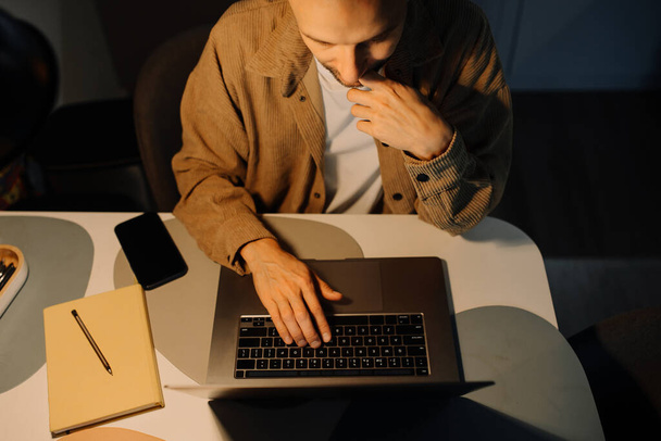 Mies työskentelee yöllä kannettavalla tietokoneella kotona pöytälampun valossa, huolissaan ja uupuneena. - Valokuva, kuva