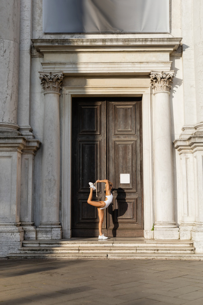 Side view of sportswoman in bodysuit and sunglasses stretching near building in Venice  - Fotó, kép