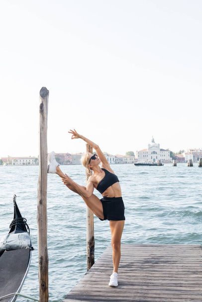 Fit sportvrouw in zonnebril, witte sneakers, zwart gewas top en korte broek stretching op pier in Venetië  - Foto, afbeelding