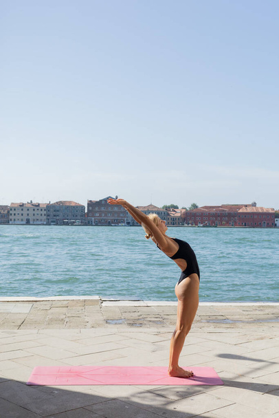 Бічний вид на біляву жінку в костюмі, що практикує йогу на набережній Венеції.  - Фото, зображення