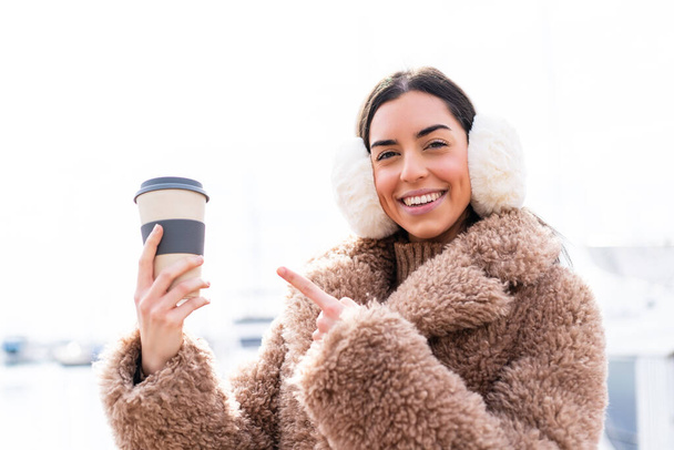 Młoda kobieta nosząca zimowe kajdanki i trzymająca kawę na zewnątrz i wskazująca ją - Zdjęcie, obraz