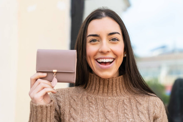 Young woman holding a wallet at outdoors with surprise and shocked facial expression - Photo, Image