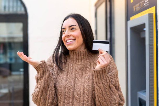 Mujer joven sosteniendo una tarjeta de crédito al aire libre con expresión facial sorpresa - Foto, Imagen