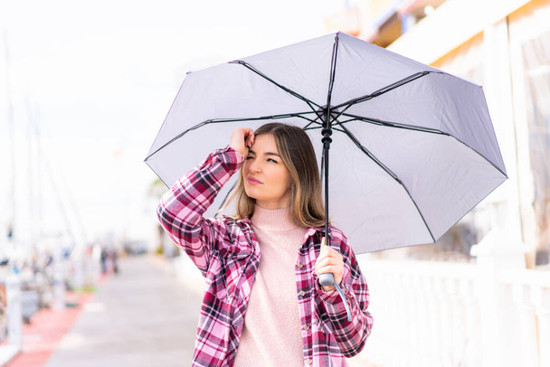 Genç, güzel Romen bir kadın şemsiyesini açık havada tutuyor, kuşkuları var ve kafası karışmış bir ifadeyle. - Fotoğraf, Görsel