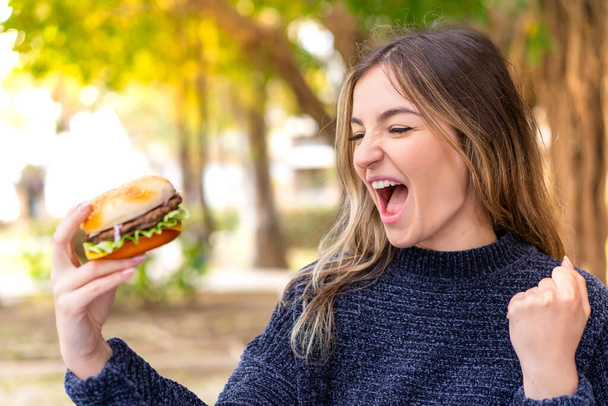 Fiatal csinos román nő kezében egy hamburger a szabadban ünnepli a győzelmet - Fotó, kép