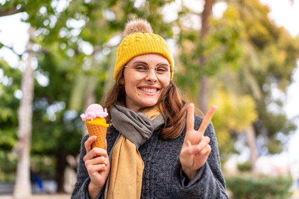 Μπρουνέτ γυναίκα με ένα cornet παγωτό σε εξωτερικούς χώρους χαμογελώντας και δείχνοντας το σημάδι της νίκης - Φωτογραφία, εικόνα