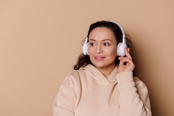 Πορτρέτο μιας όμορφης πολυεθνικής γυναίκας με μπεζ κουκούλα, ακούγοντας μουσική σε ασύρματα ακουστικά και χαμογελώντας κοιτάζοντας στην άκρη στο χώρο της διαφήμισης αντιγράφων, σε απομονωμένο φόντο κρέμας. Έννοια ψυχαγωγίας - Φωτογραφία, εικόνα