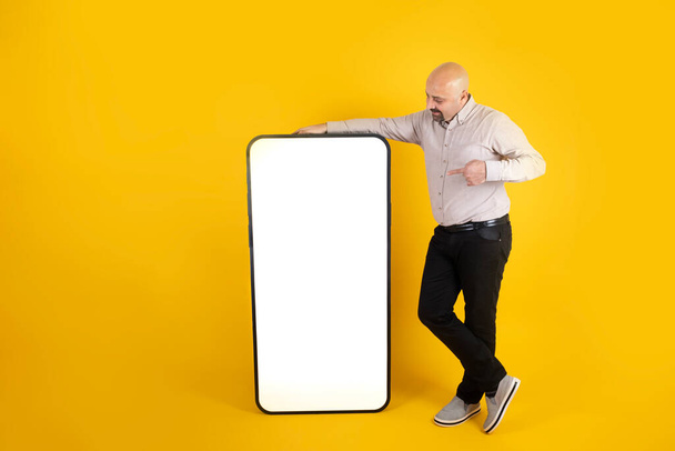 Big blank smartphone, full body length of man pointing finger at big blank smartphone. Human sized mobile phone mock up. Yellow background. Smiling guy showing empty white screen. Ad concept idea. - Фото, зображення