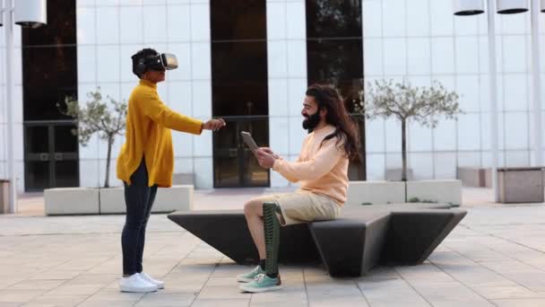 Молодая пара использует очки VR в центре города с наушниками. Красивая латиноамериканка пробует виртуальную реальность 360 видео и 3D видео приложение человек с протезом ноги использует пульт дистанционного управления - Кадры, видео