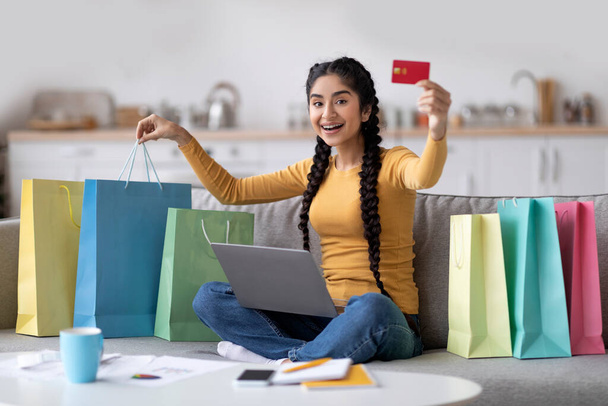 Online Shopping. Happy Young Indian Woman Using Laptop And Showing Credit Card At Camera While Sitting On Couch At Home Next To Bright Shopper Bags, Smiling Hindu Female Enjoying Internet Purchases - 写真・画像