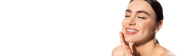 pleased young woman with natural makeup and bare shoulders touching cheek isolated on white, banner  - Foto, Imagen