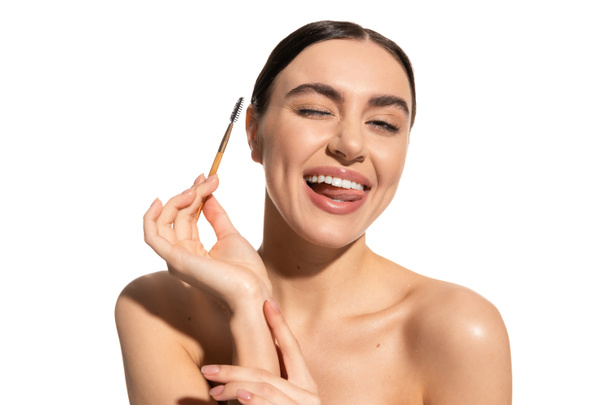 happy young woman sticking out tongue while holding eyebrow brush isolated on white  - 写真・画像