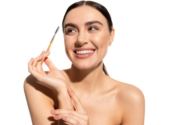pleased young woman with bare shoulders holding eyebrow brush isolated on white  - Φωτογραφία, εικόνα