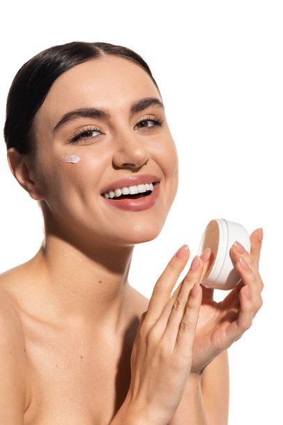 joyful woman with bare shoulders holding jar with moisturizing face cream isolated on white  - Photo, image