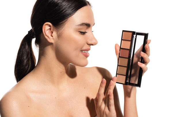 positive young woman with bare shoulders holding eye shadow palette isolated on white  - Photo, Image