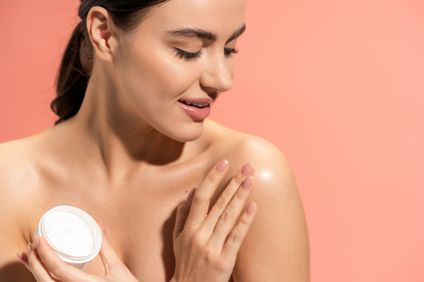 portrait of cheerful woman holding jar while applying nourishing cream on bare shoulder isolated on pink  - Fotoğraf, Görsel