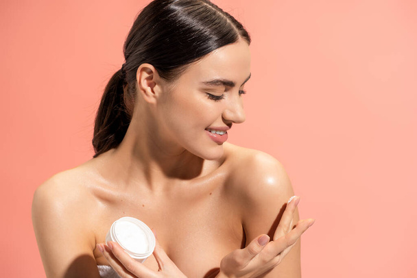 smiling woman holding jar and applying nourishing cream on bare shoulder isolated on pink  - Foto, afbeelding