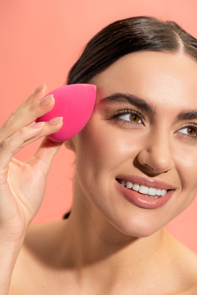 χαρούμενη νεαρή γυναίκα εφαρμογή μακιγιάζ ίδρυμα με σφουγγάρι ομορφιάς απομονώνονται σε ροζ  - Φωτογραφία, εικόνα