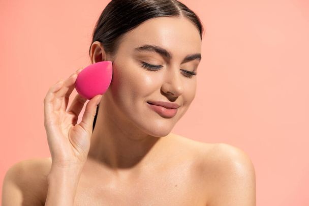 pleased woman applying face foundation with makeup sponge isolated on pink  - Zdjęcie, obraz