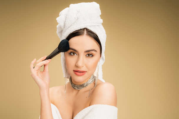 charming woman with white towel on head applying face powder with makeup brush on beige background  - Fotografie, Obrázek