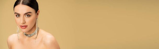 brunetta giovane donna in collana e orecchini guardando la fotocamera isolata sul beige, banner  - Foto, immagini