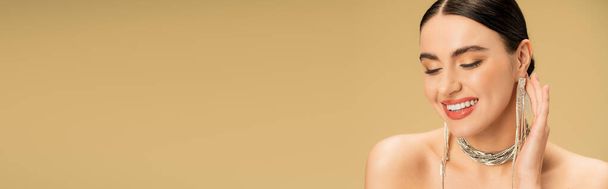 brunette young woman in necklace and earrings smiling isolated on beige, banner  - 写真・画像