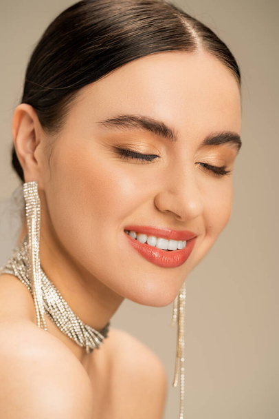 joyful young woman in necklace and earrings smiling with closed eyes isolated on beige  - Foto, Bild