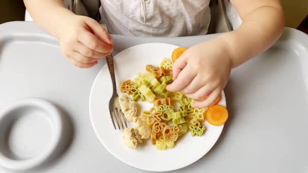 Cute toddler baby kid boy enjoy having fun sit in chair learning eat with fork,hands,plate healthy carrot vegetables,animal shape pasta at home kitchen. Children healthy food nutrition diet concept. - Materiaali, video