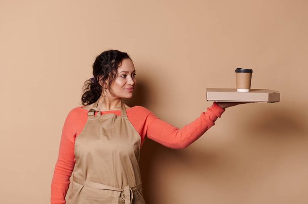 Attractive African American woman waitress, barkeeper, wearing beige apron, holding a cardboard flat box with tasty Italian pizza and takeaway hot drink in eco paper cup, isolated on cream background - Photo, Image