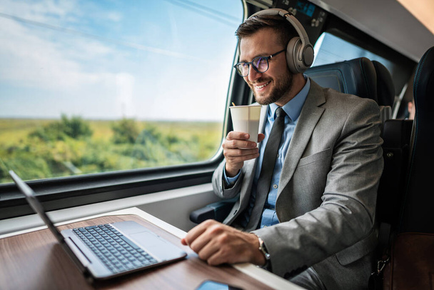 Handsome businessman is having a good time while traveling by high-speed train. He is using laptop computer and wireless headphones for online communication, gaming and entertainment. - Valokuva, kuva