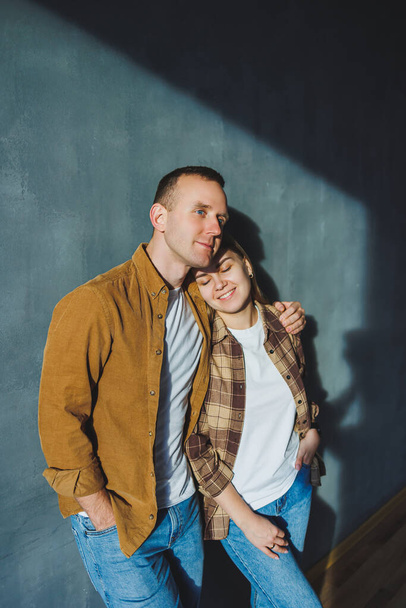 A young married couple in love in shirts and jeans on the background of a gray wall. The concept of happy family relationships. A man and a woman are hugging - Fotó, kép