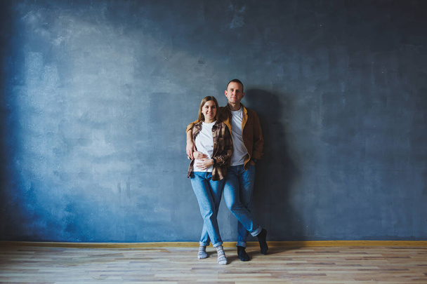 A young married couple in love in shirts and jeans on the background of a gray wall. The concept of happy family relationships. A man and a woman are hugging - Фото, изображение
