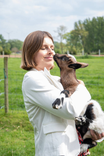 Young woman plays with goat kids, feeding them, sun shining over farm in background, High quality photo - Photo, Image