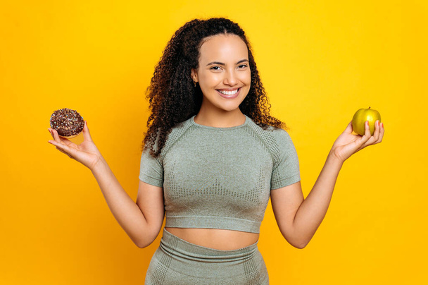 Positive lovely brazilian or hispanic curly haired sporty woman, in sports outfit, holds fresh apple and sweet donut, makes a choice, stand on isolated orange background, looks at camera, smiling - Photo, Image