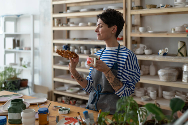 Artisan tattooed girl drink coffee and looking at tasty bun lustily in art studio during break from producing handmade pottery for handicraft shop. Potter woman has lunch with tea on workplace - Photo, Image