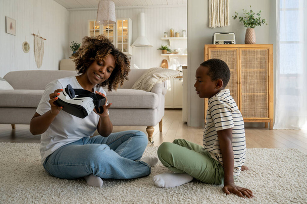 Joven madre afroamericana sentada en el suelo con hijo niño sosteniendo gafas de realidad virtual. Madre negra padre explicando la tecnología VR al niño. Regalos de alta tecnología para niños - Foto, Imagen