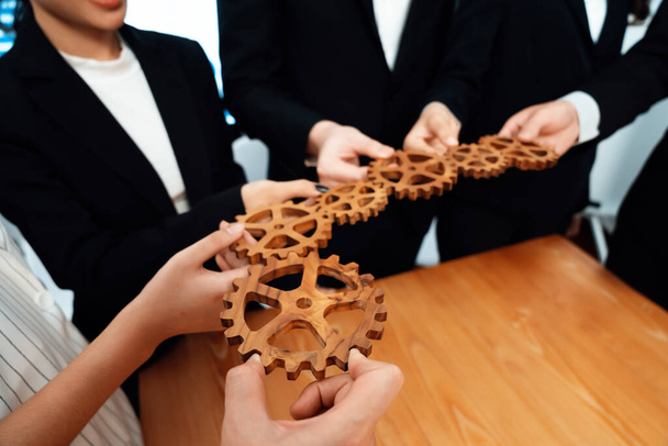 Closeup hand holding wooden gear by businesspeople wearing suit for harmony synergy in office workplace concept. Group of people hand making chain of gears into collective form for unity symbol. - Фото, изображение