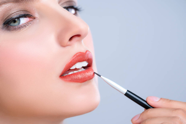 Close-up mooie jonge vrouw met vlekkeloze gezonde huid en natuurlijke make-up zetten verleidelijke mode glanzende rode lippenstift op haar lip met lip borstel in geïsoleerde achtergrond. - Foto, afbeelding