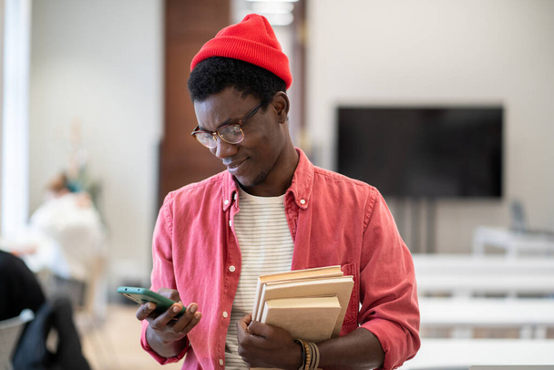 Pleasant zwarte student man staat in de campus bibliotheek met leerboeken met behulp van smartphone op studiepauze. Tevreden Afro-Amerikaanse man scroll webberichten, chat online, surfen op internet in de klas - Foto, afbeelding