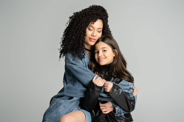 Curly woman in denim jacket hugging happy daughter isolated on grey  - Foto, Imagem