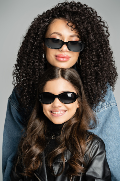 Portrait of curly woman in sunglasses posing near smiling daughter isolated on grey  - Zdjęcie, obraz
