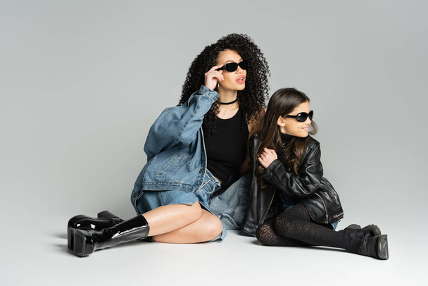 Trendy woman and happy daughter in sunglasses sitting on grey background  - Zdjęcie, obraz