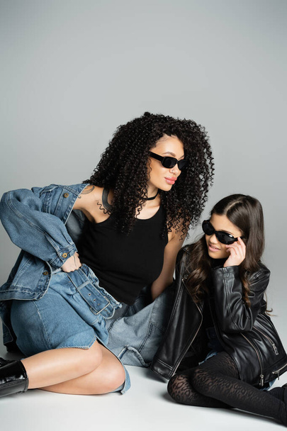 Stylische Frau in Jeansjacke und Sonnenbrille sitzt neben Tochter auf grauem Hintergrund  - Foto, Bild