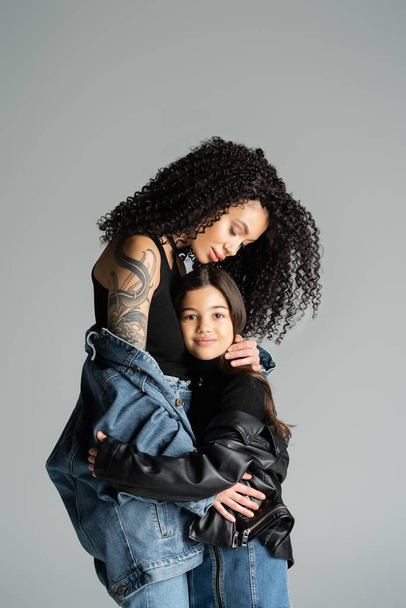 Tattooed mother in denim jacket hugging daughter isolated on grey  - Фото, изображение